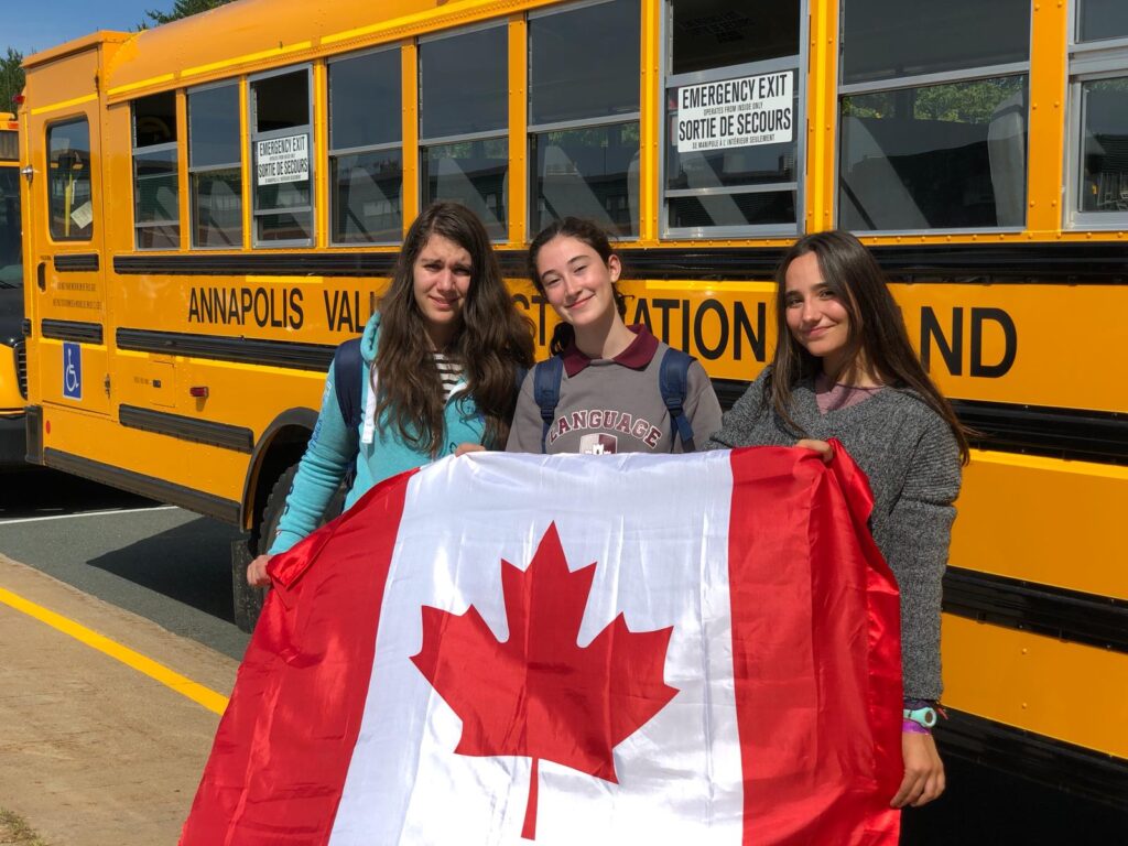 Tre studentesse con la bandiera del Canada davanti a uno scuolabus giallo.