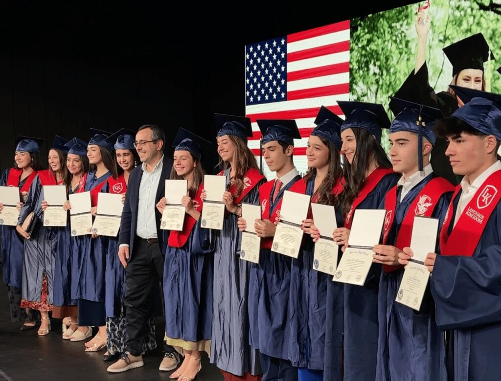 Gruppo di studenti sul palco mostra l’US High School Diploma davanti alla bandiera degli Stati Uniti.