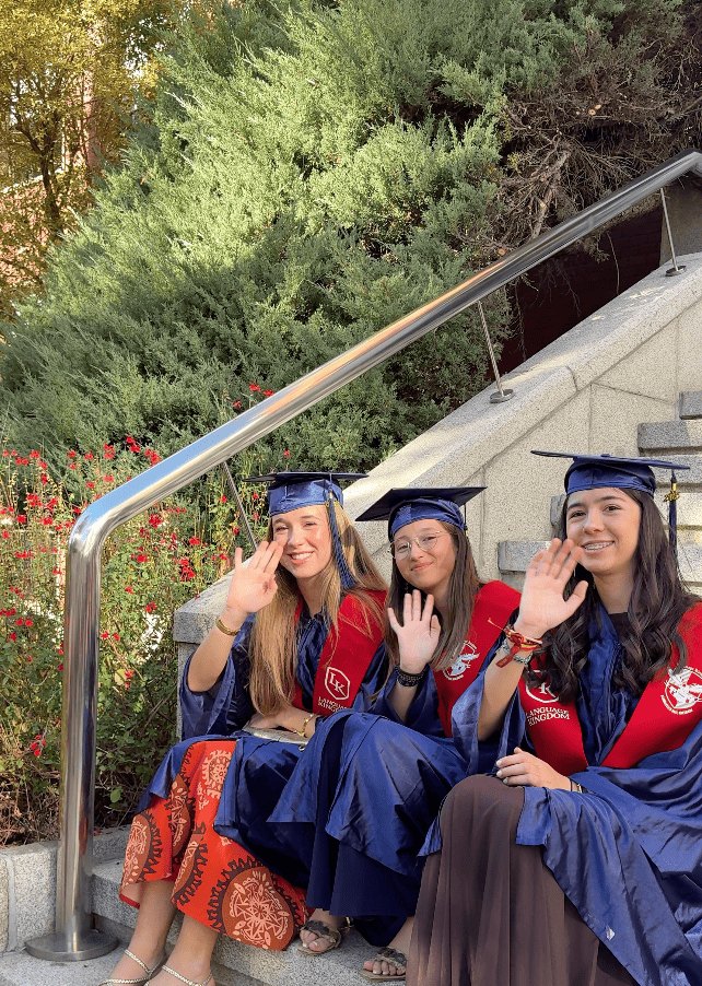 Tre studentesse con toga blu e stola rossa salutano sedute su una scalinata il giorno della graduation.